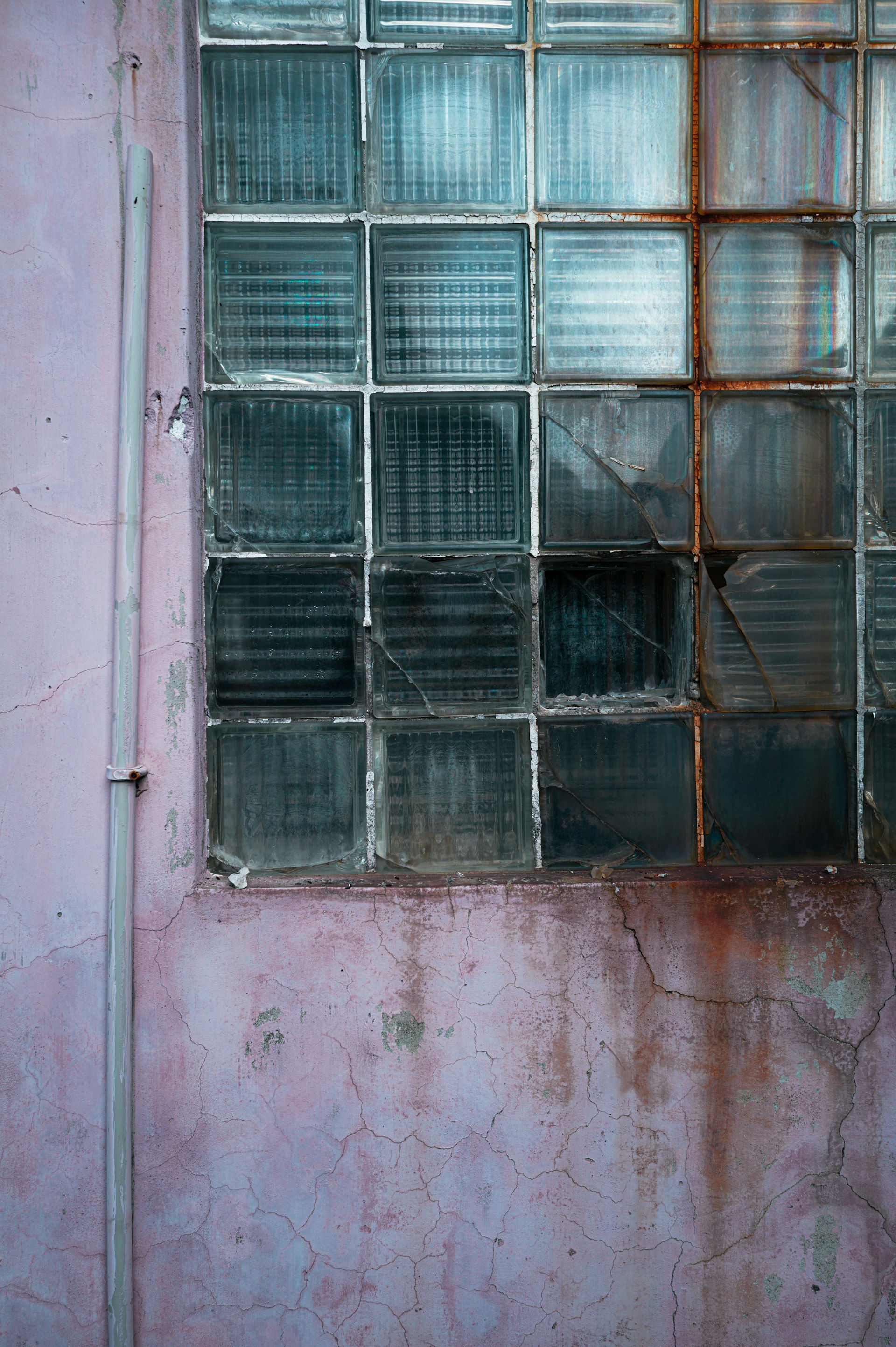 Weathered glass block window set into a cracked pink wall, glass still catching light despite decay
